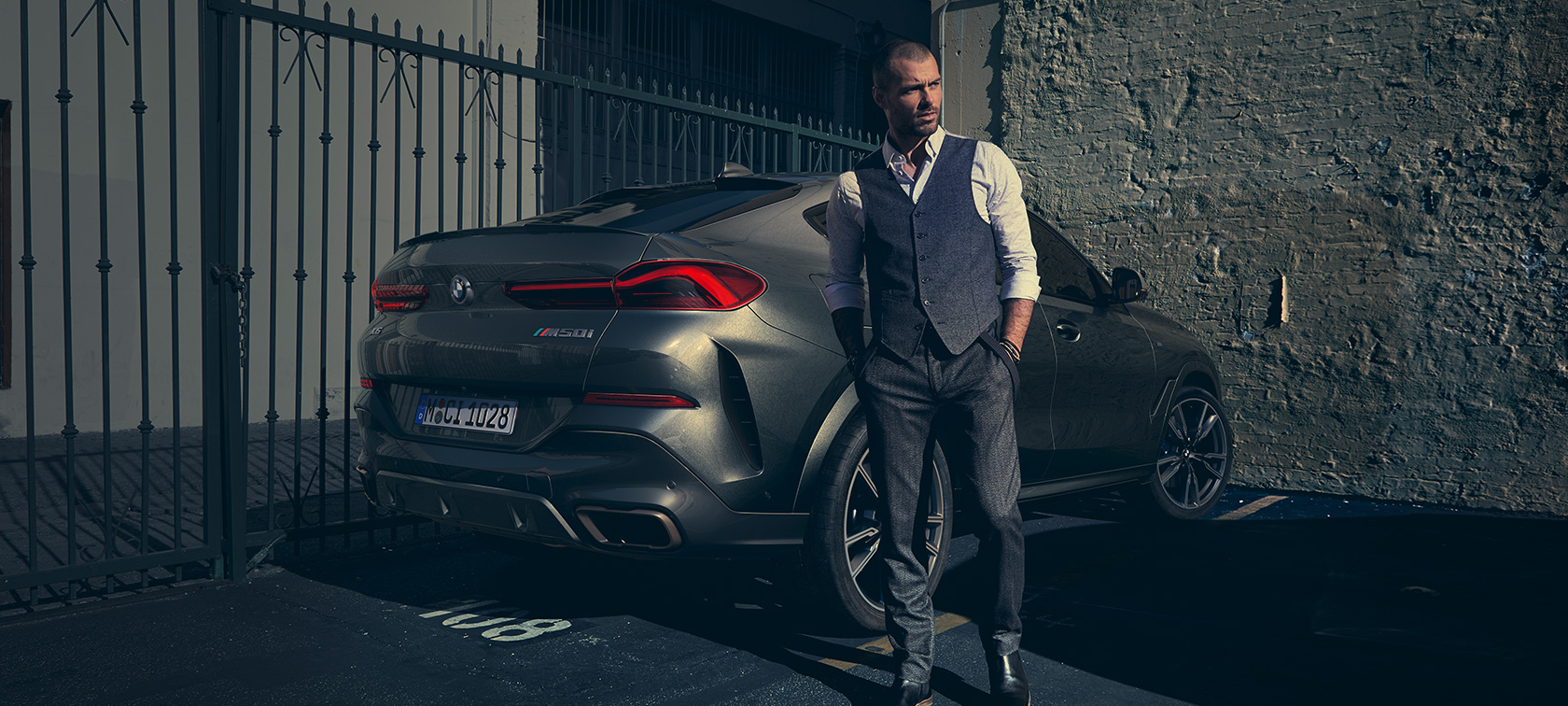 BMW X6 M50i, вид сзади с поворотом в три четверти. Мужчина стоит рядом с BMW X6 M50i, вид сзади с поворотом в три четверти.