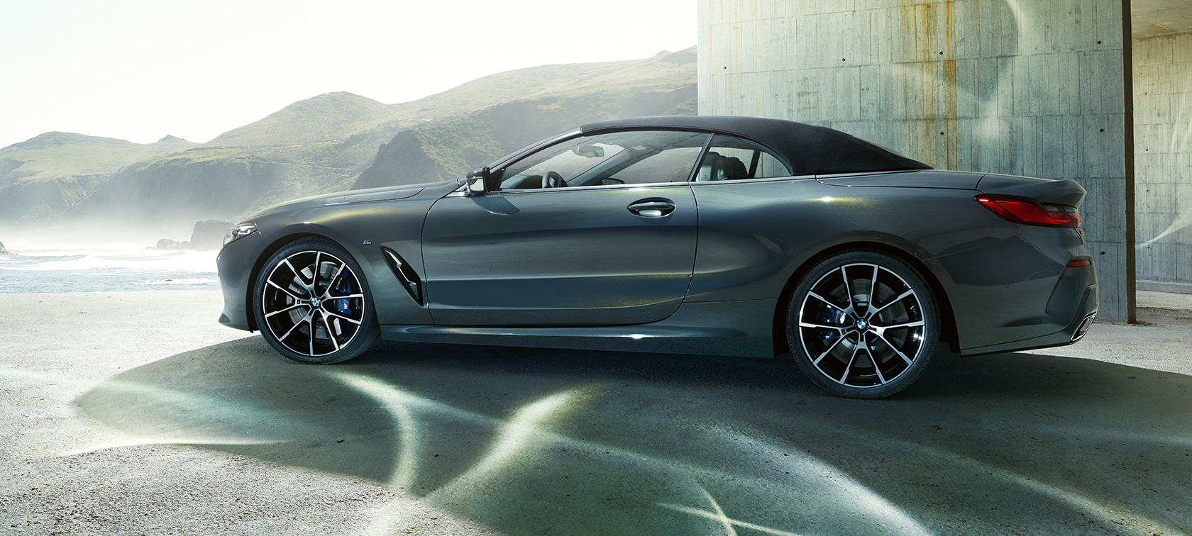 BMW M850i xDrive Кабриолет, вид сбоку. BMW M850i xDrive Кабриолет в цвете Dravit Grey металлик с закрытым мягким верхом, вид сбоку перед холмами.