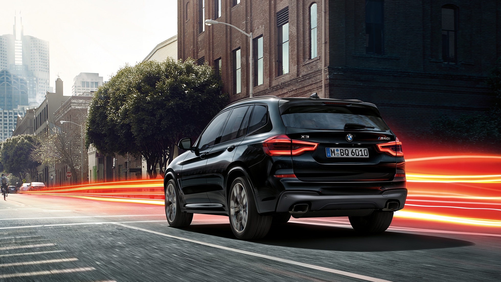 BMW X3 M40i, вид сзади с поворотом в три четверти. BMW X3 M40i в Black Sapphire металлик, за рулем, экстерьер, вид сзади с поворотом в три четверти на городском фоне со световыми полосками.